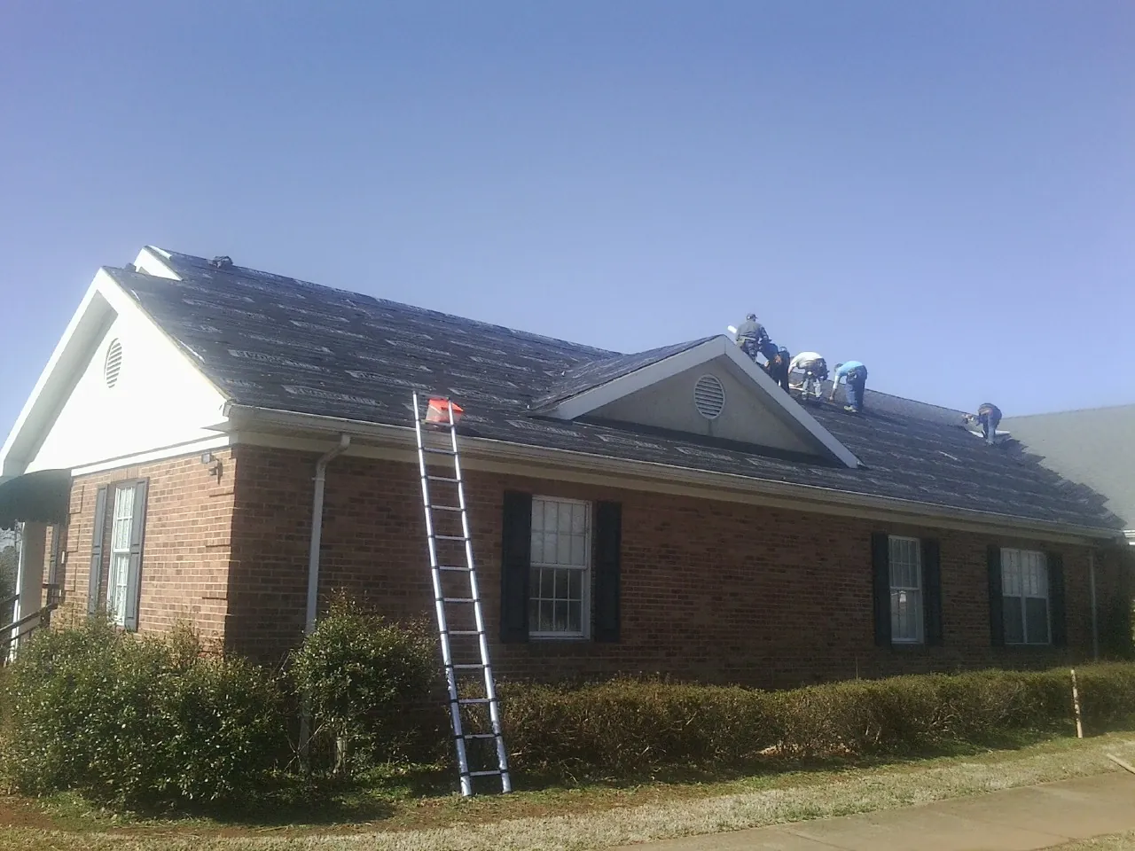Crew tearing shingles off a brick ranch roof with a ladder set — Metal Roof Repair work by our crew in Dyersville