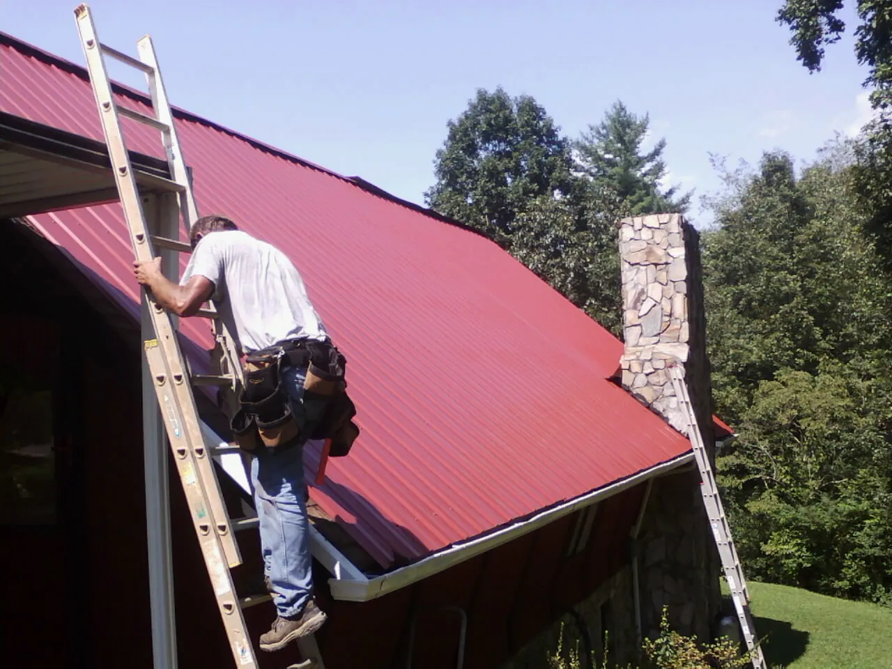 Roofer on a ladder beside a red metal roof and stone chimney — Soffit & Fascia Repair by Roofing Brothers in Dyersville